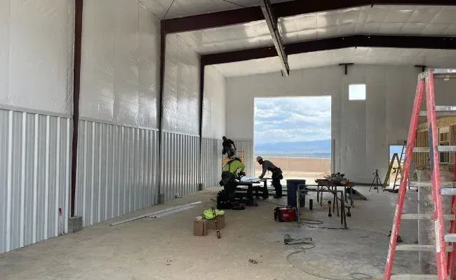 Two construction workers in a large, unfinished metal warehouse frame, with an open bay door leading to a field.