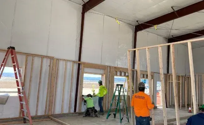 Construction workers building wooden stud walls inside a warehouse with white walls and a red ladder.