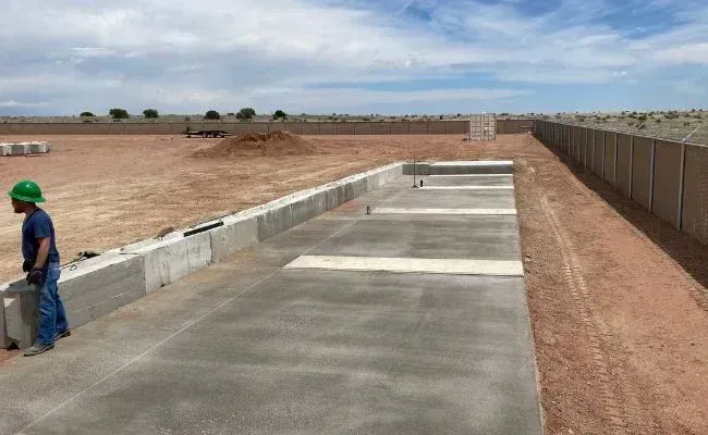 A construction worker in a hard hat stands near a long, newly poured concrete foundation on a flat, dusty site.