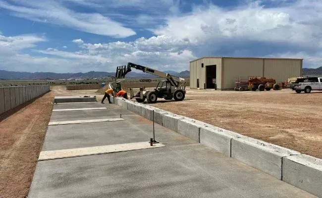 Workers in high-visibility gear use a telehandler to install concrete barrier blocks along a paved outdoor construction site.