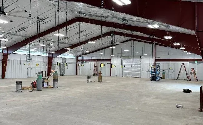 A large, empty industrial warehouse with a high, white ceiling, metal support beams, and scattered construction equipment.
