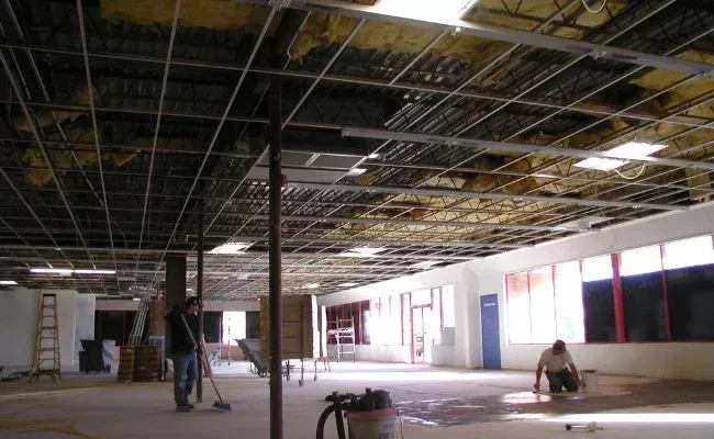 Two workers renovate a commercial space with exposed ceiling grids, unfinished walls, and scattered construction equipment.