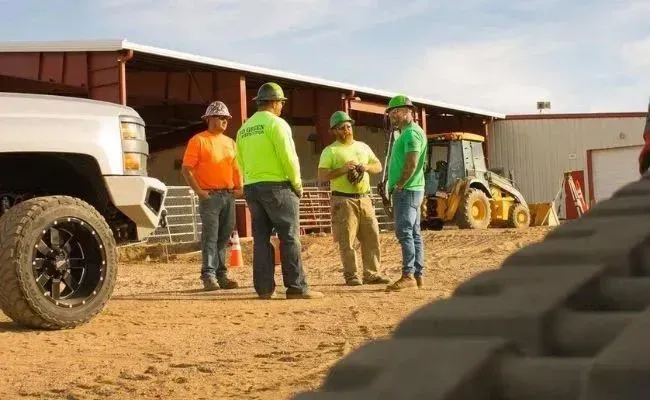 Four construction workers in high-visibility gear talk on a dirt job site near a truck and heavy equipment.