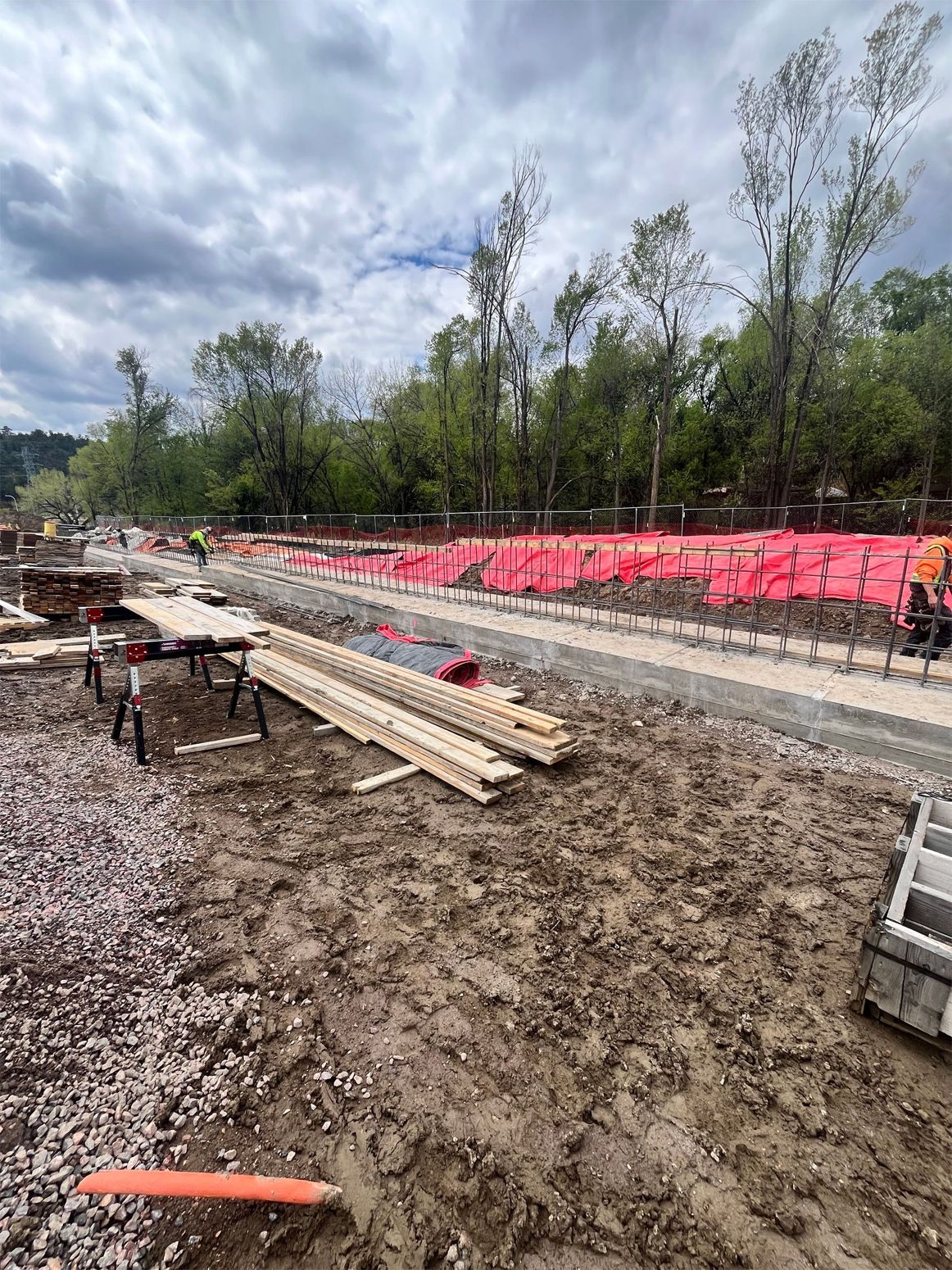 Construction site with muddy ground, stacked lumber, orange safety fencing, and cloudy sky
