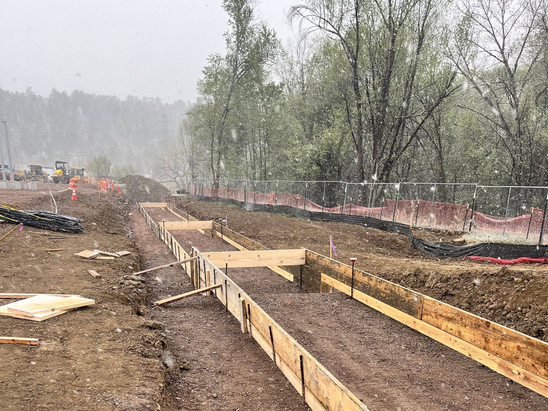 Construction site with rebar foundation forms and muddy ground beside a tree-lined road in light rain