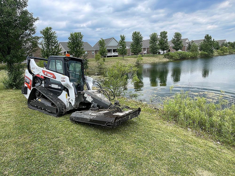 A bobcat is cutting grass next to a lake.