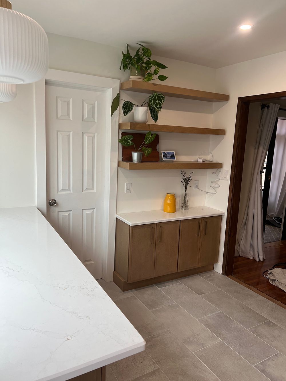 Bright kitchen nook with open shelves, brown cabinet, white door, and countertop by a tiled floor.