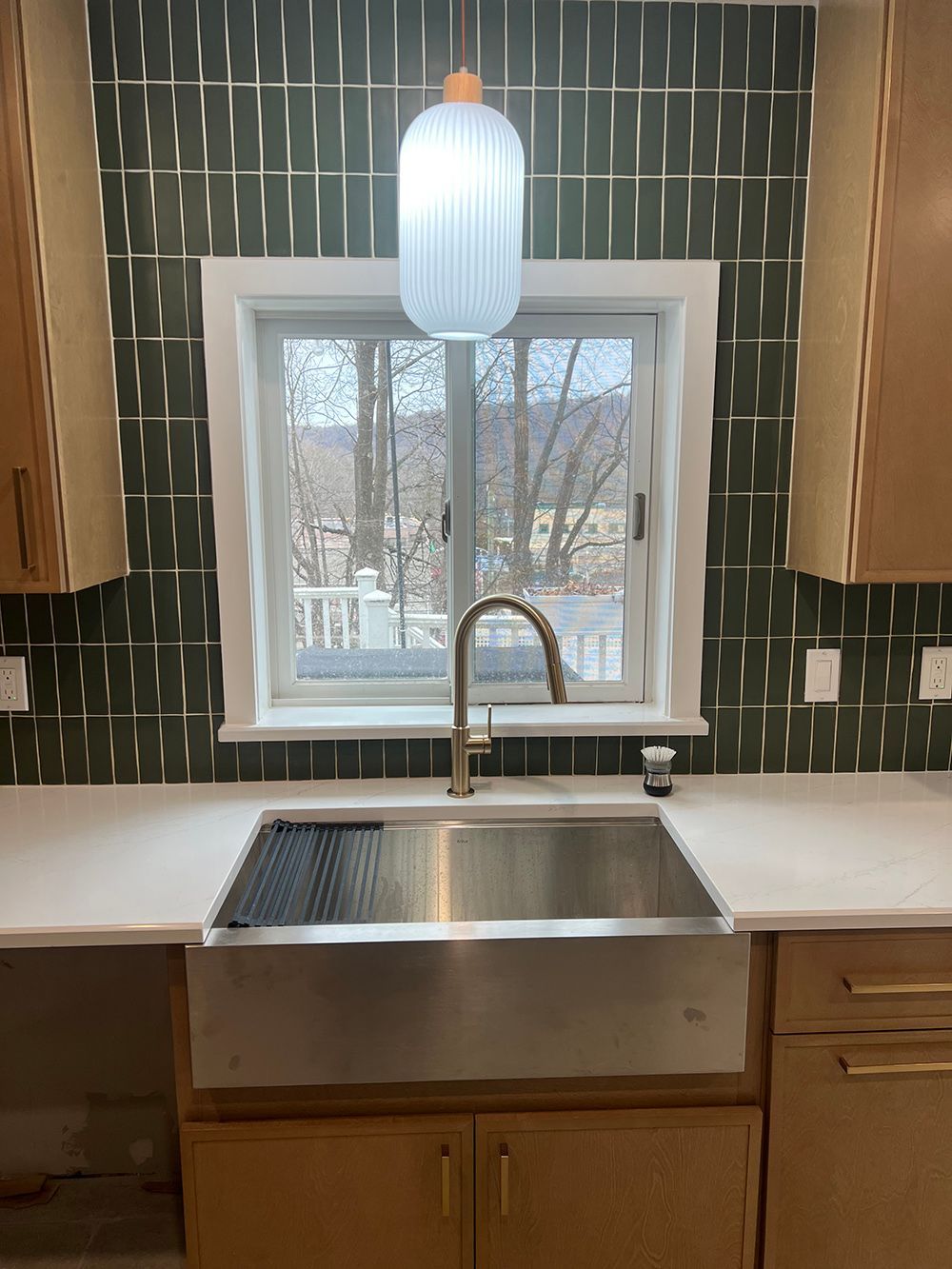 Kitchen sink under window with green tile backsplash, white pendant light, and wooden cabinets