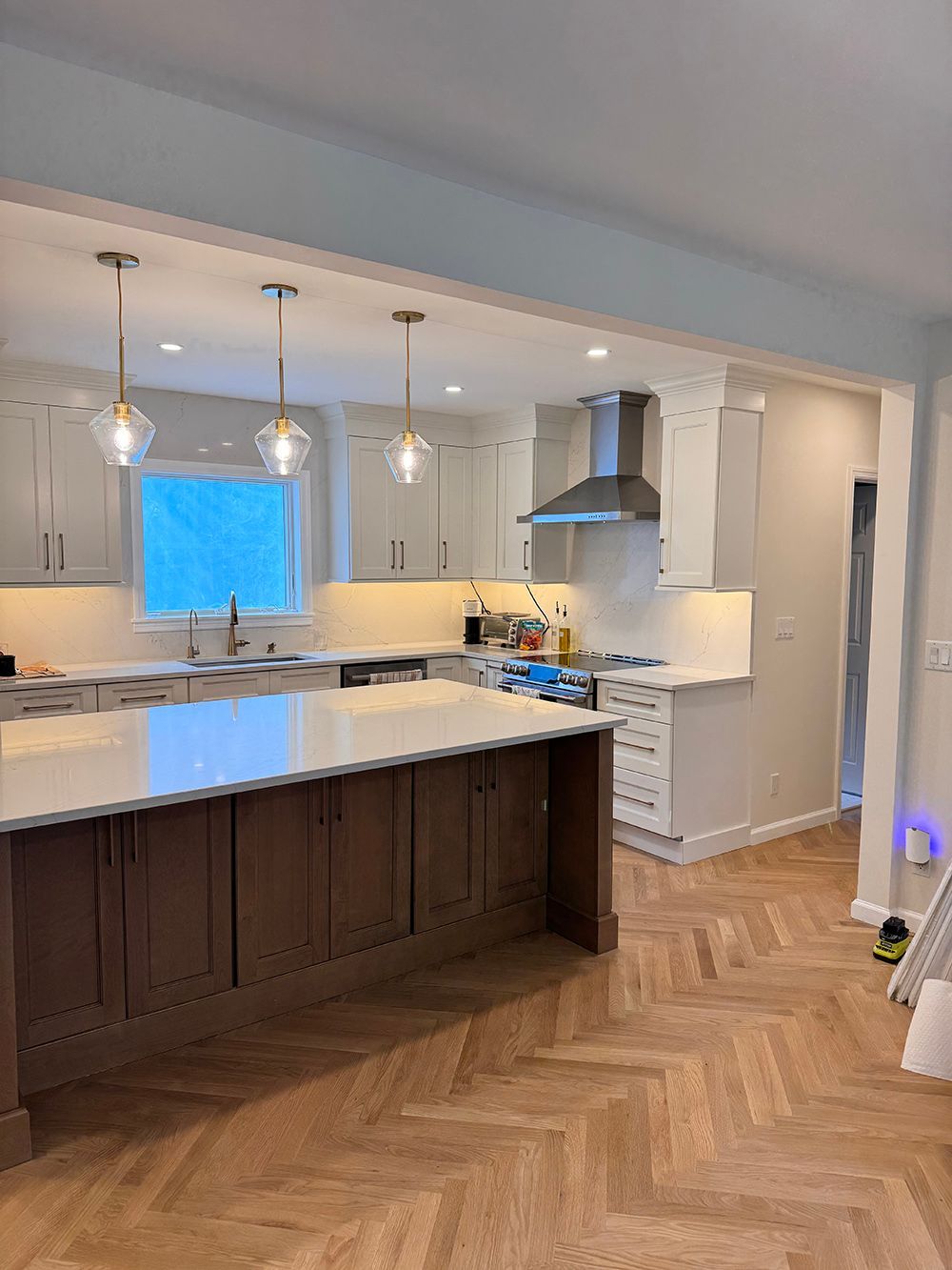 Bright modern kitchen with white cabinets, dark island, pendant lights, and herringbone wood floor.
