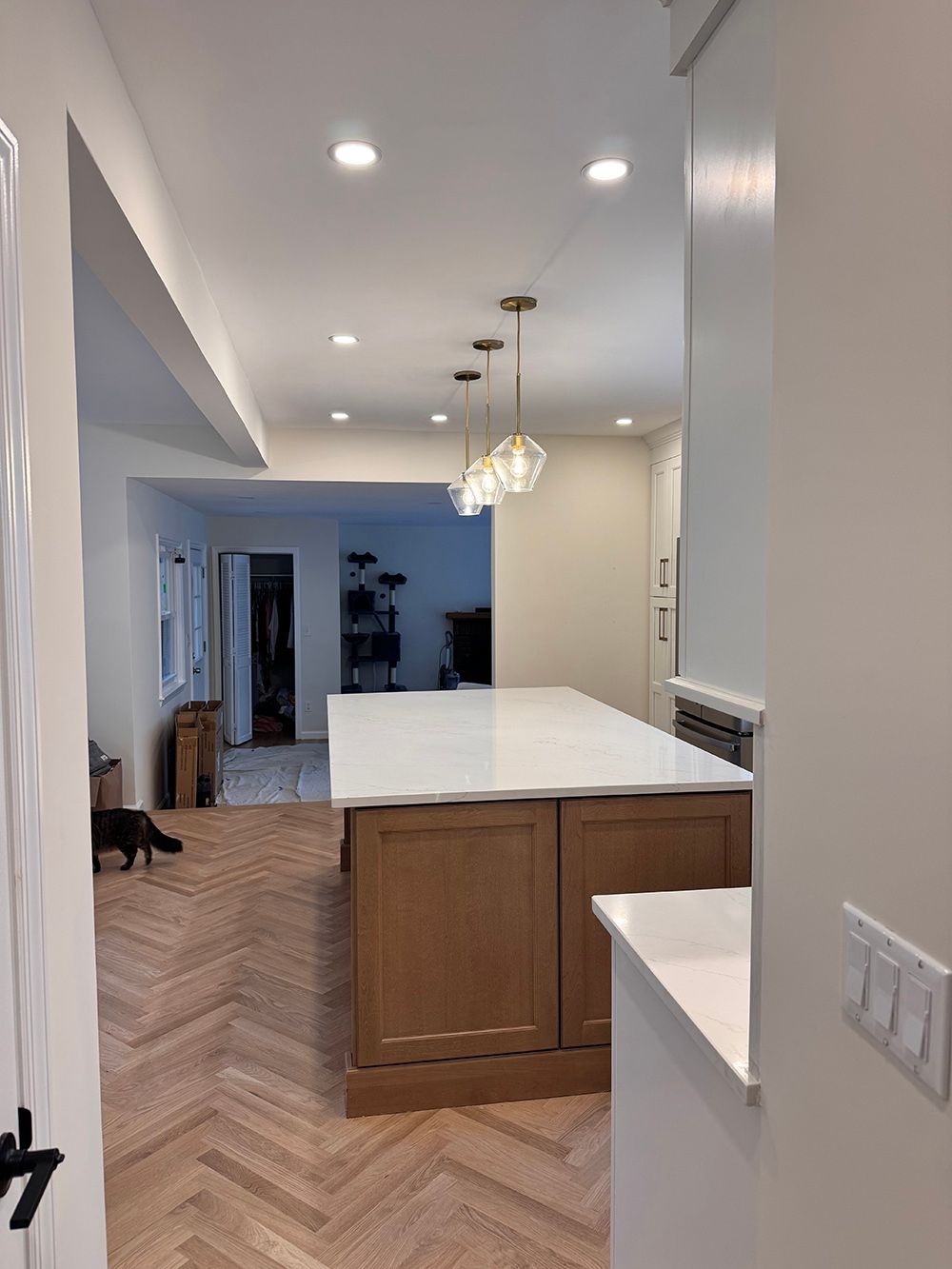 Bright kitchen with white island, wood cabinets, herringbone floor, and recessed ceiling lights opening to a hallway