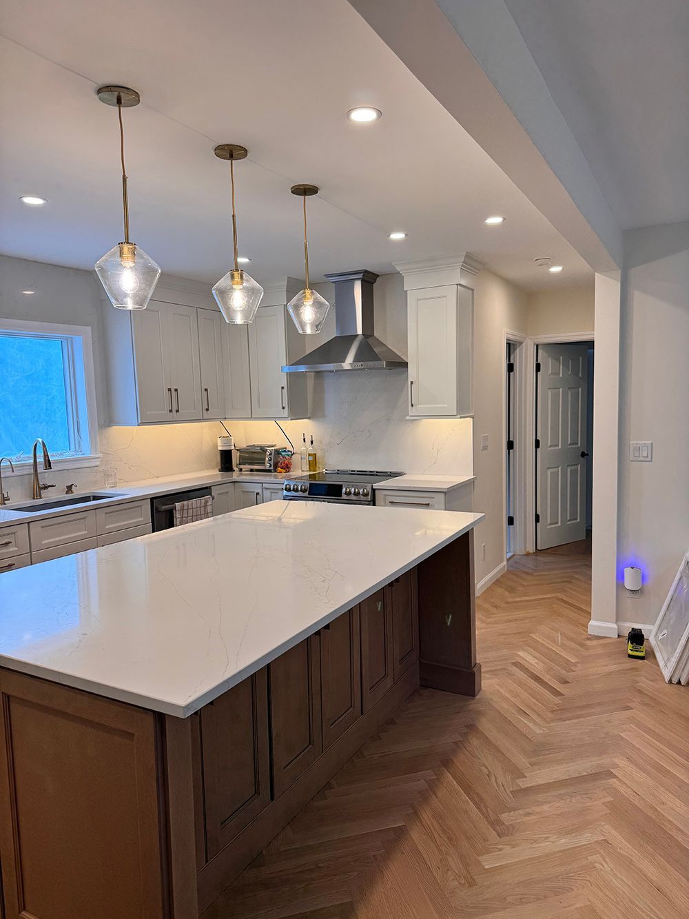 Modern kitchen with white island, pendant lights, herringbone wood floor, and stainless steel appliances