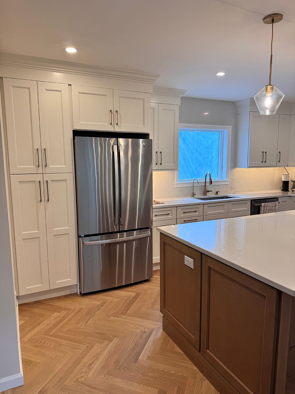 Bright kitchen with stainless steel fridge, white cabinets, and brown island in herringbone wood floor.