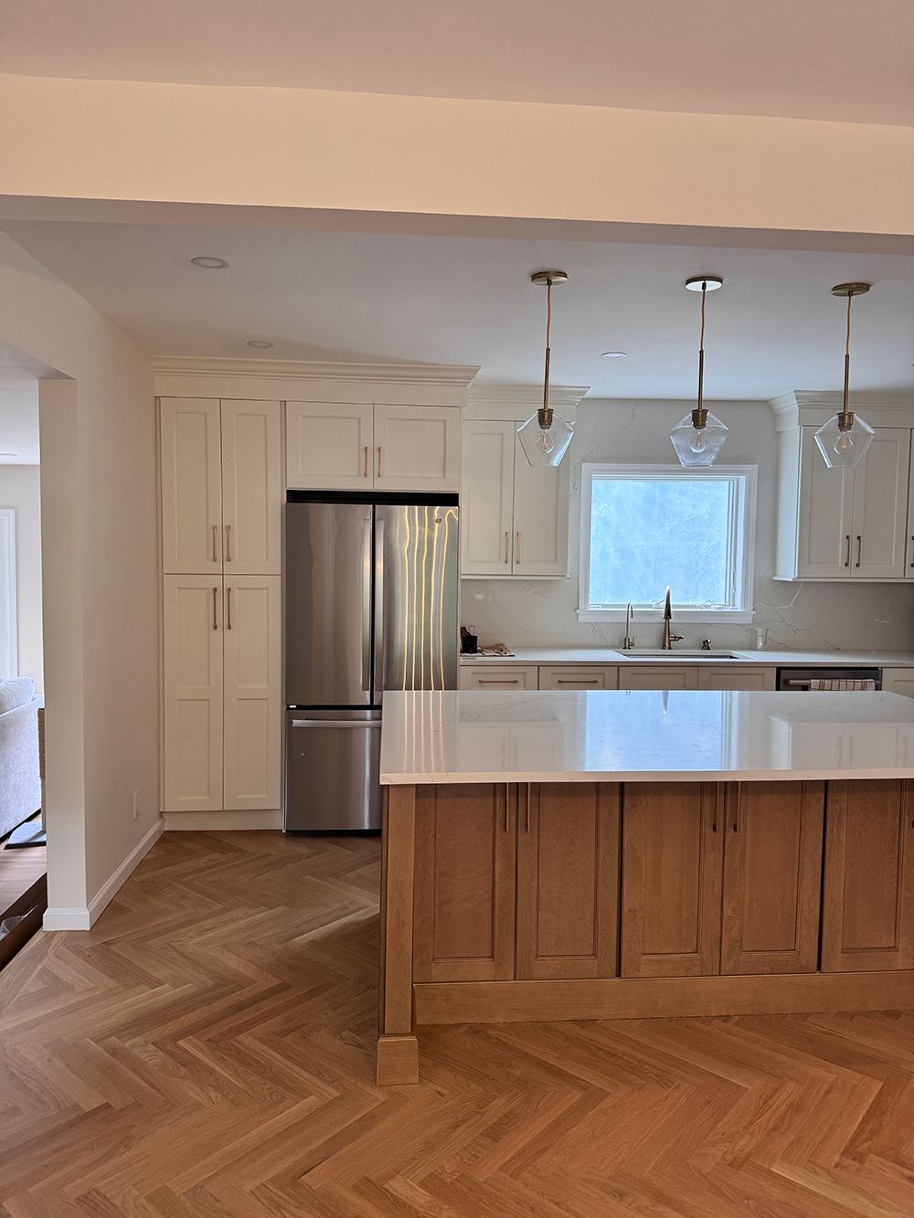 Bright modern kitchen with stainless fridge, white island, herringbone wood floor, and window over sink