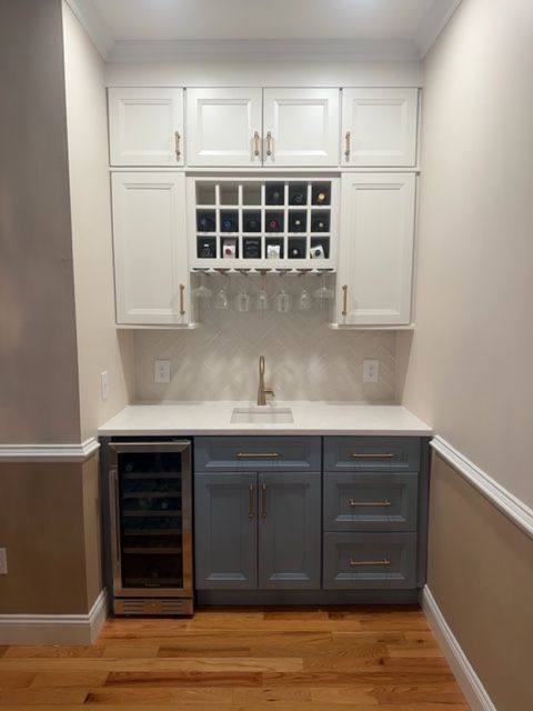 Compact home bar with white cabinets, gray lower cabinets, wine rack, and wood floor.