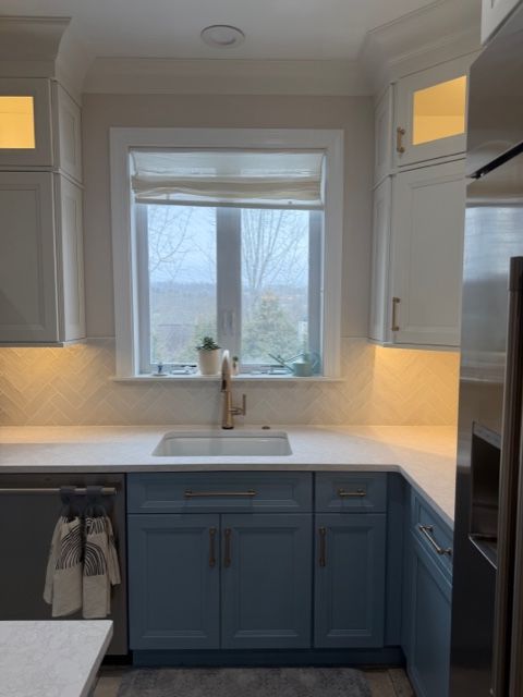Kitchen sink under a window with blue cabinets and white countertops, lit by warm under-cabinet lights.
