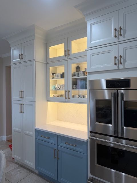 Built-in kitchen cabinets with lit glass shelves and stainless steel refrigerator beside a blue lower cabinet.