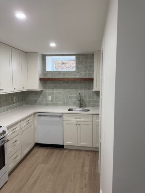Compact white kitchen with gray backsplash, wood floor, sink, dishwasher, and small window