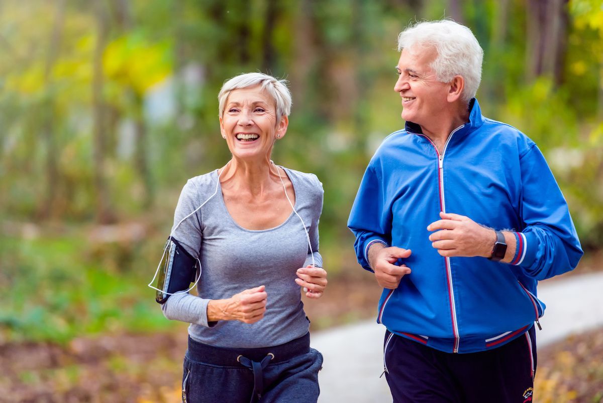 Smiling couple jogging on a path in a park; woman wearing grey, man wearing blue.