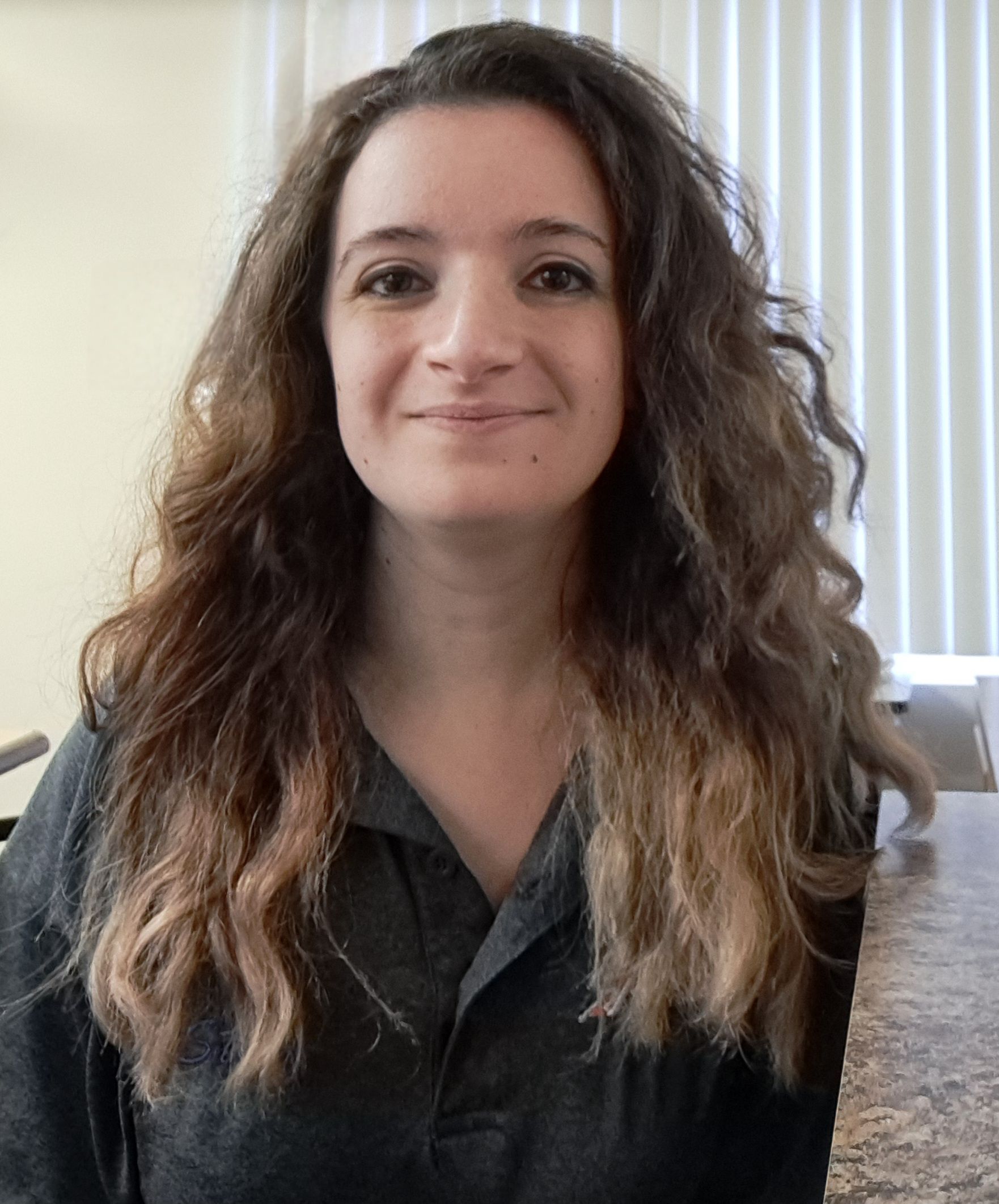 Woman with long, wavy brown hair and a slight smile, wearing a dark shirt, indoors near a window.