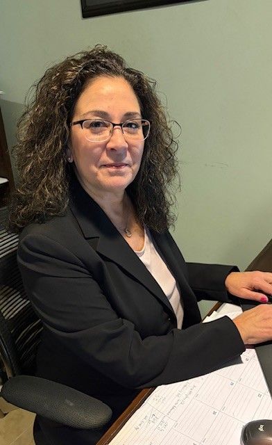 Woman with glasses smiling, dark hair, wearing a black top, outdoors in front of a building.