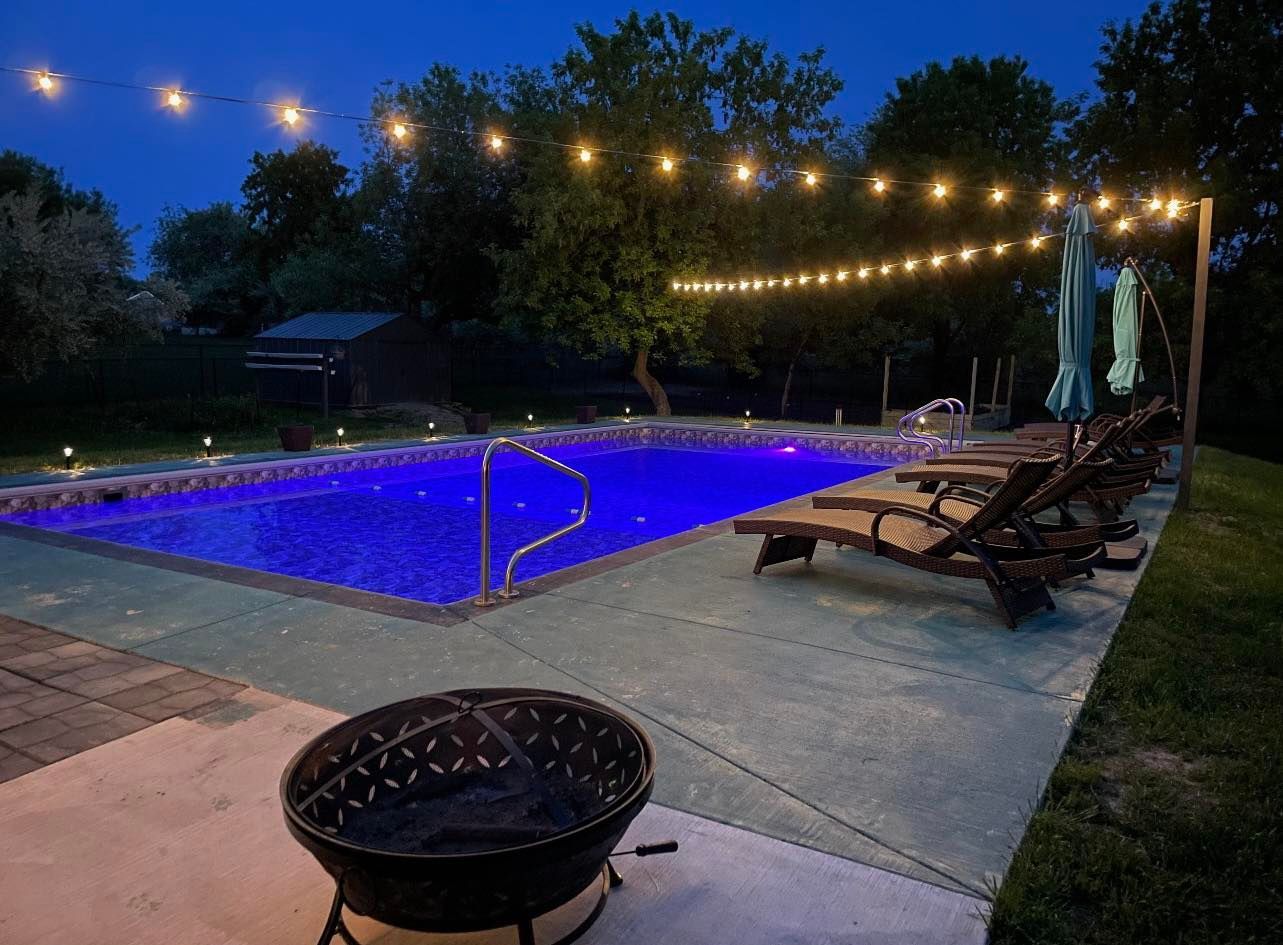 A large swimming pool is lit up at night with a fire pit in the foreground.