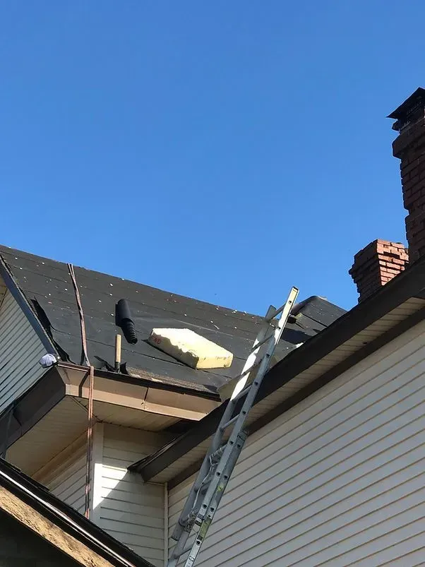 Ladder leaning on a house roof; a partially replaced section is visible, with a brick chimney and blue sky background.