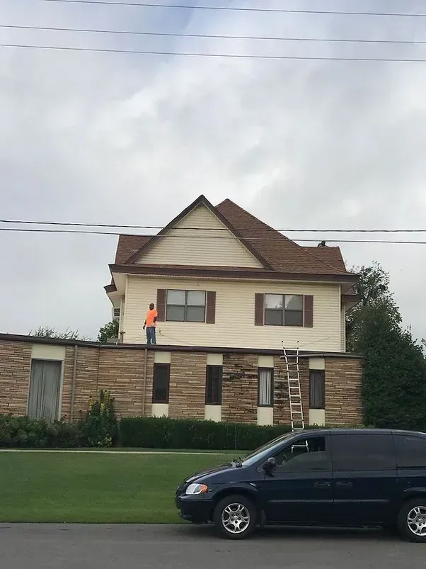 Person painting upper level of two-story house, brown roof, tan siding, ladder, with van parked in front.
