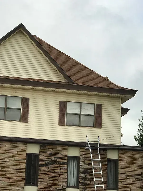 Two-story house with brown roof and trim, tan siding, and brick veneer, with a ladder against the wall.