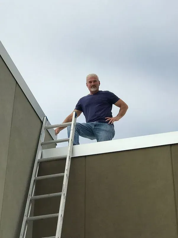 Man on a roof, crouched near a ladder, looking at the camera. Overcast sky.
