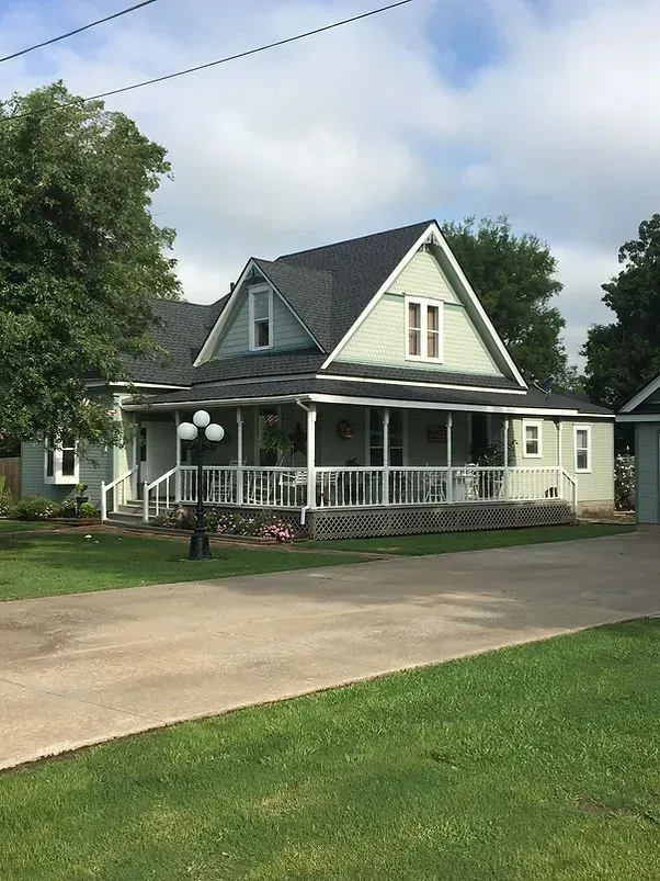 Green house with wrap-around porch, dark roof, on a grassy lawn.