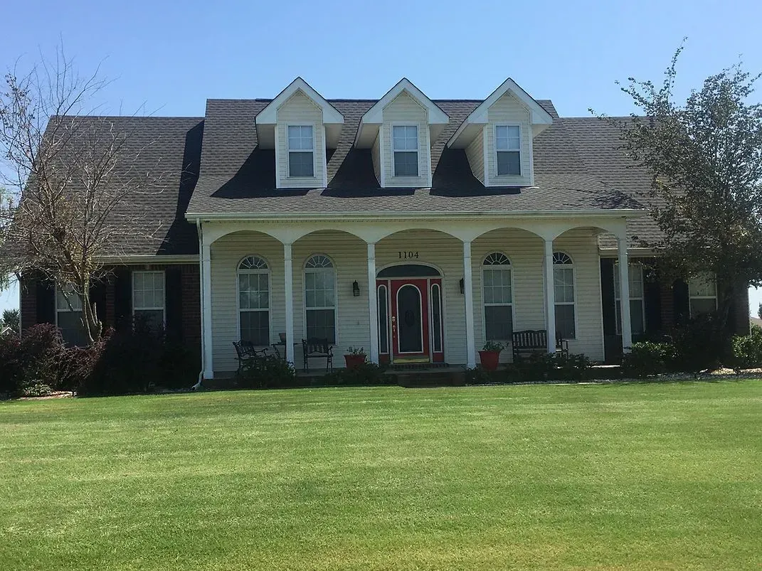 A light-colored house with a porch, dormers, and a large green lawn under a blue sky.