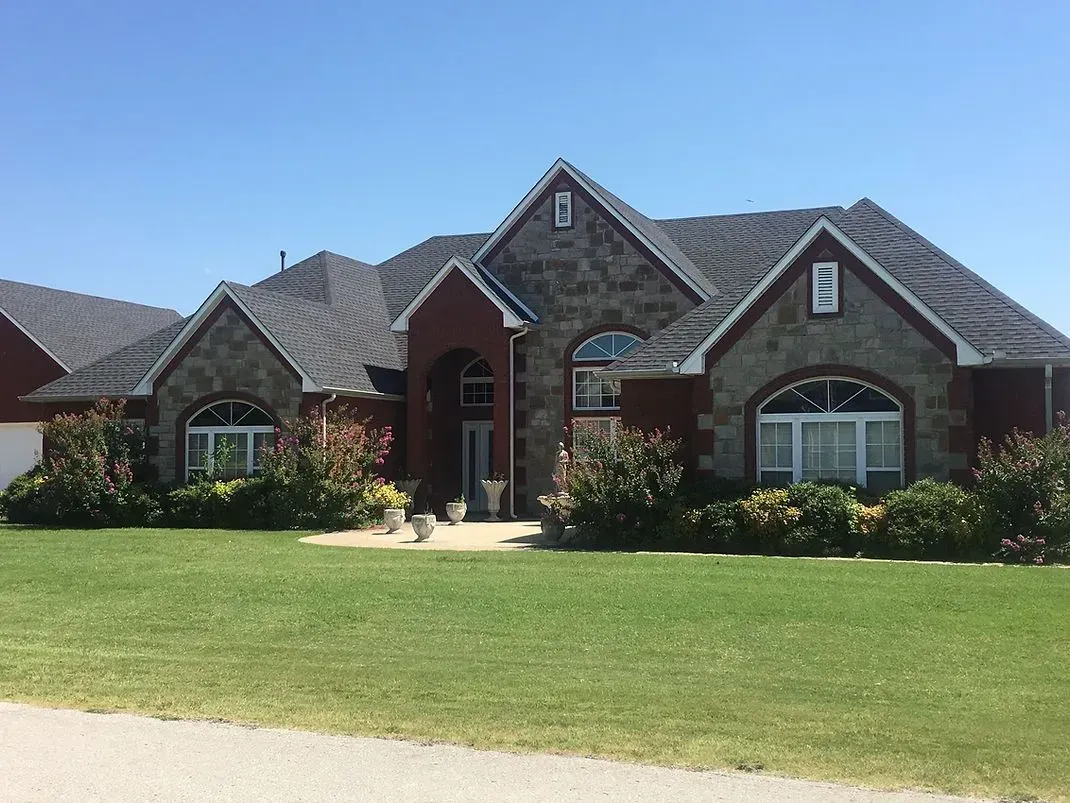 House with stone and red brick facade, arched windows, and lush landscaping on a sunny day.