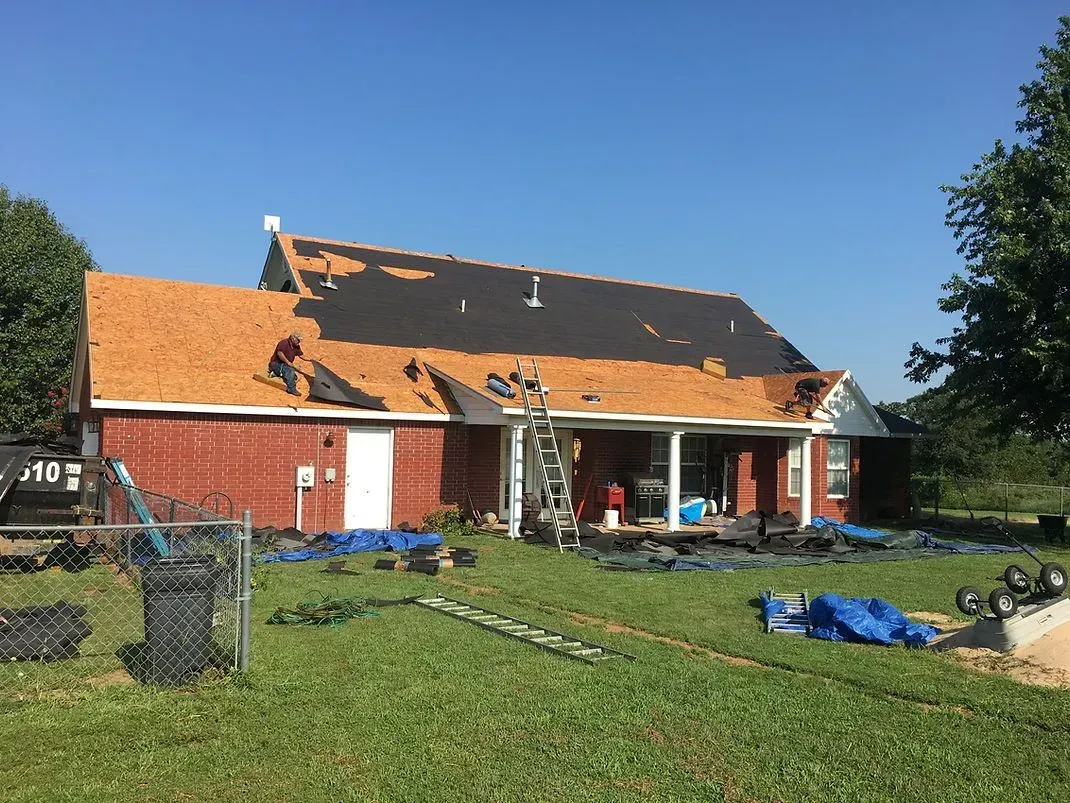 A brick house has roof partially removed, workers on roof, surrounded by tarps, tools, and a ladder.