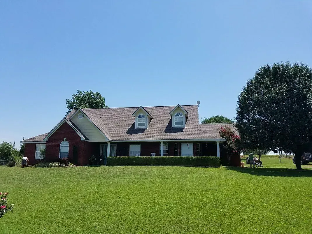 Brick house with a covered porch, three dormer windows, and a large tree on a sunny day.