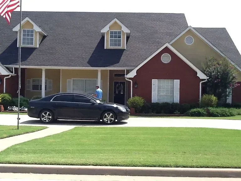 Black car parked in front of a large house with a green lawn and American flag.