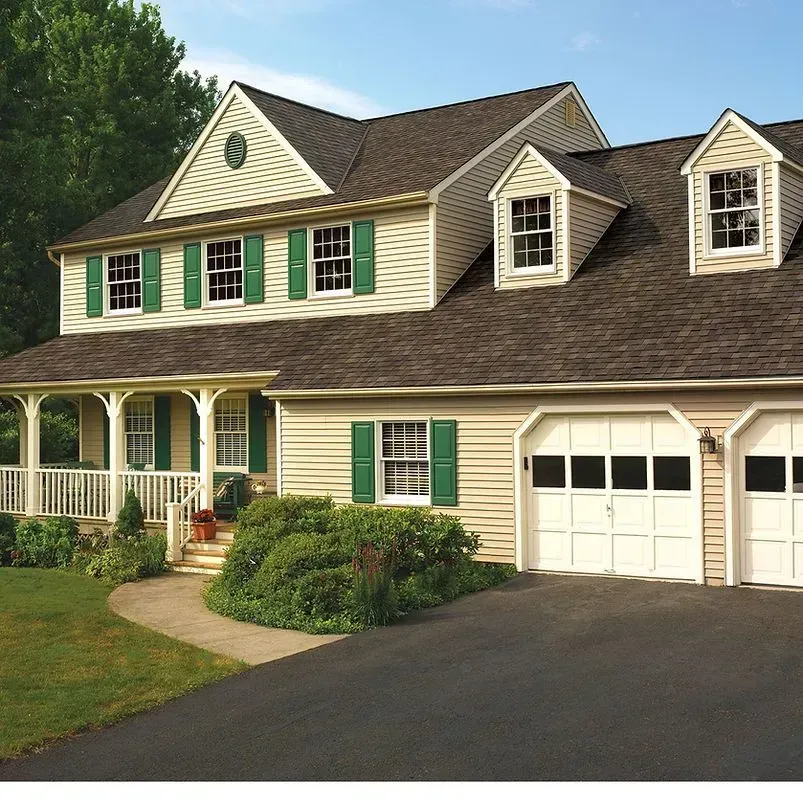Beige house with green shutters, white garage doors, and a brown roof. Sunny day.