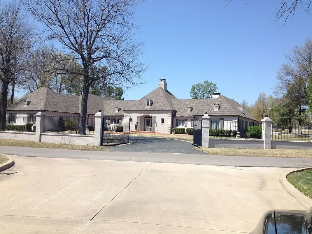 A large, light-colored house with a multi-gabled roof and open gate. Trees and blue sky in the background.