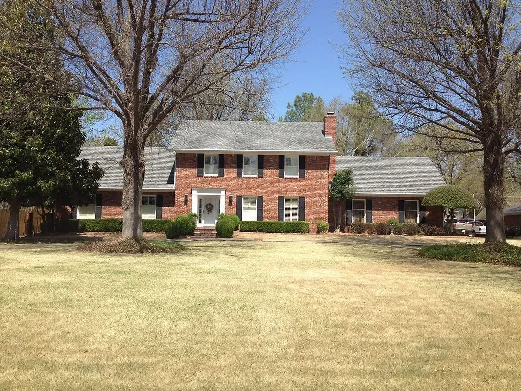 Red brick house with gray roof, green lawn, and bare trees against a blue sky.