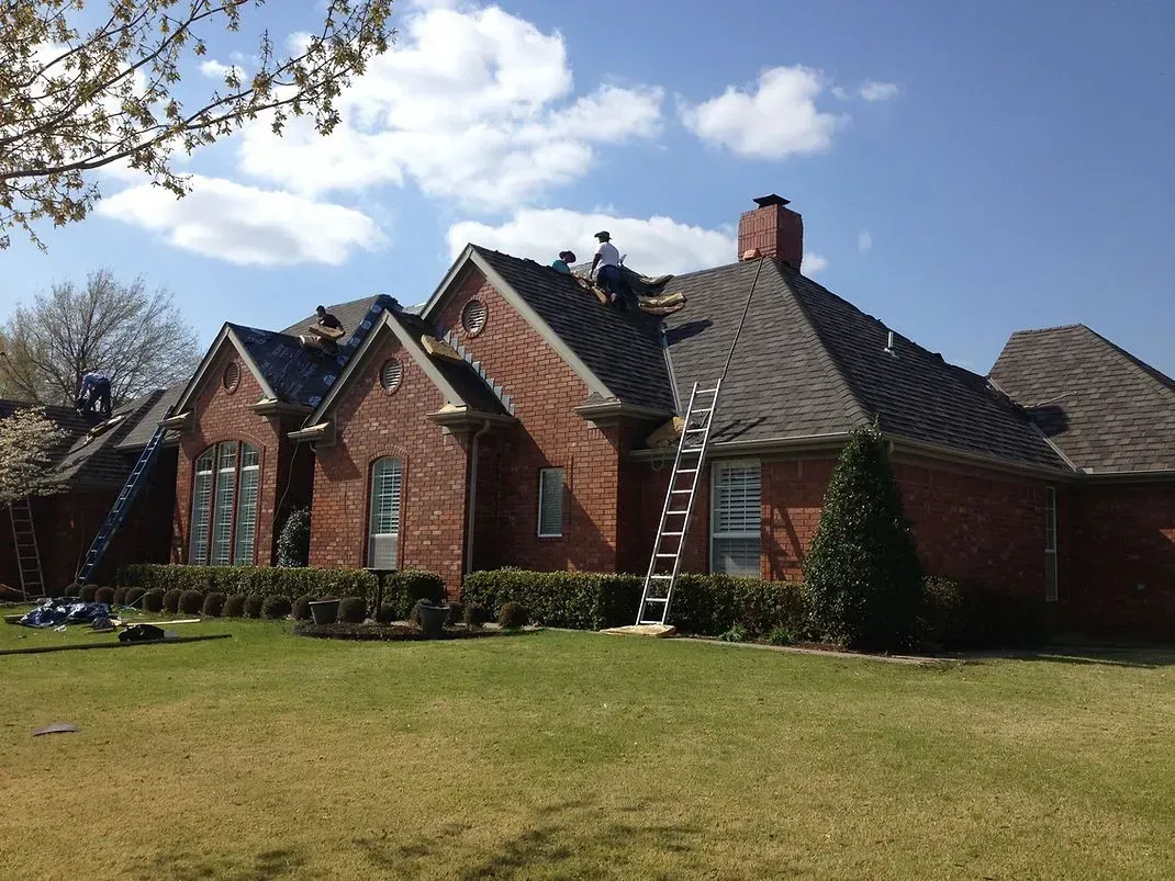 Roofers on a brick house replacing shingles; ladders, blue sky, and green grass visible.