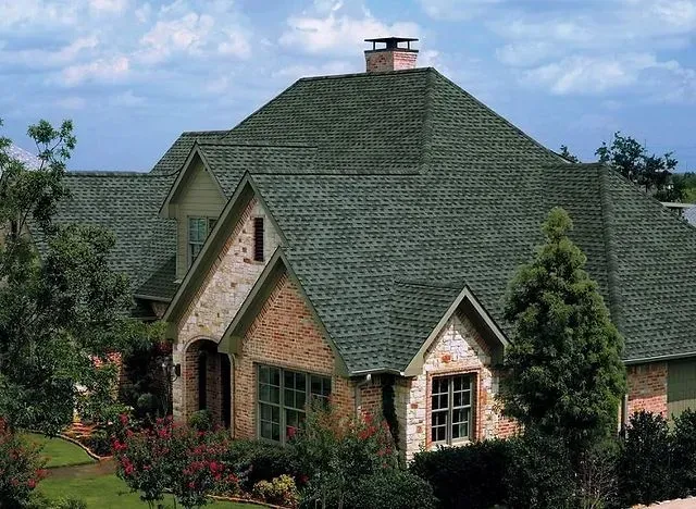 A house with a green roof, brick and stone exterior, windows, and surrounding greenery.