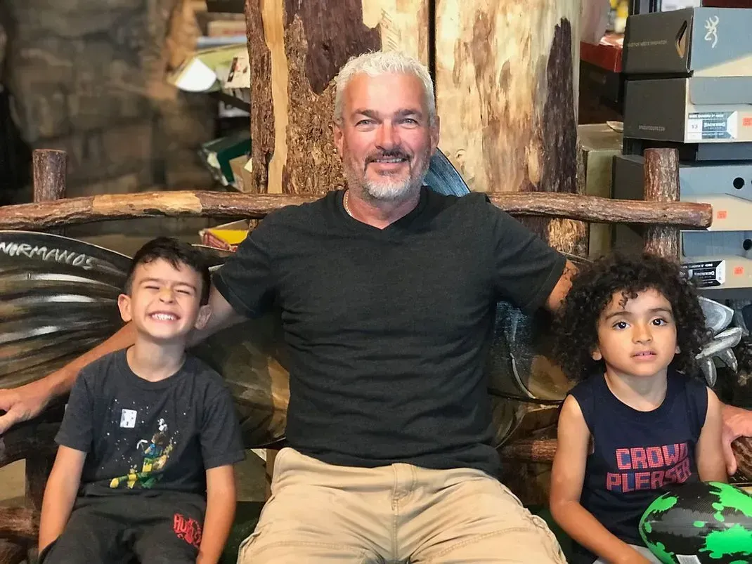 Man with graying hair sits between two smiling young boys; rustic chair.