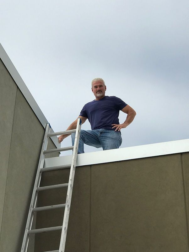 Man on rooftop, climbing a ladder. Blue shirt, jeans. Cloudy sky.