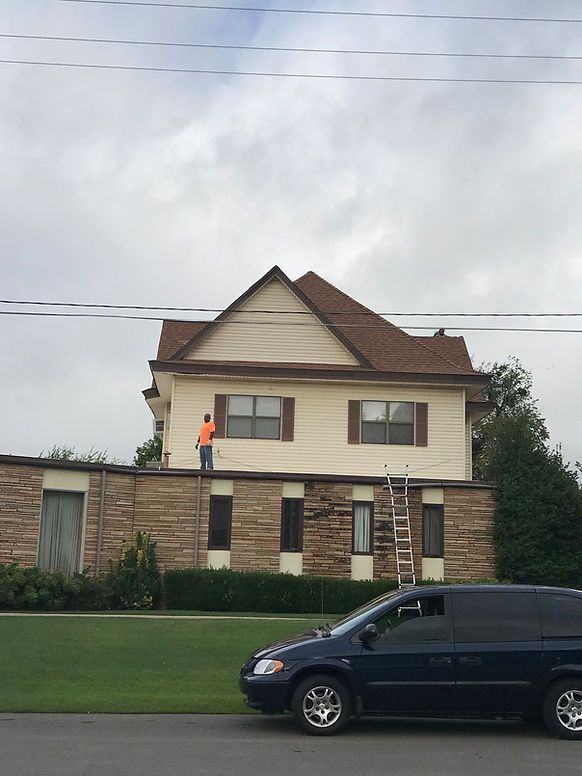 Person painting trim on two-story house with brown roof and stone facade, parked blue van in front. Cloudy sky.