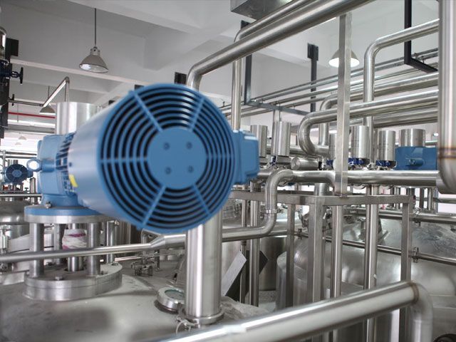 Blue industrial motor connected to stainless steel pipes and tanks in a factory setting.