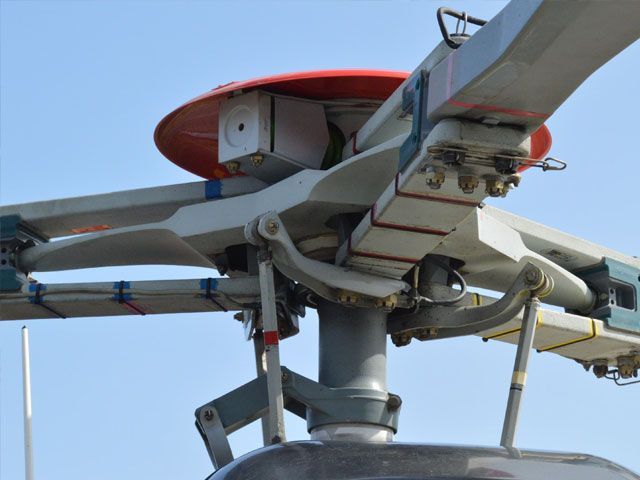 Helicopter rotor system with red top, grey blades, and metal components. Blue sky in the background.
