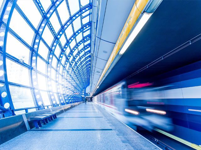 Blue subway station with a passing train, modern architecture, and a bright sky visible through large windows.