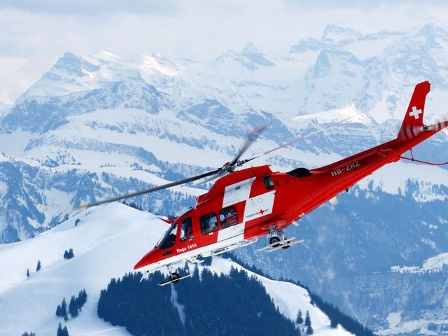 Red and white helicopter flying over snowy mountains, Swiss flag on the tail.