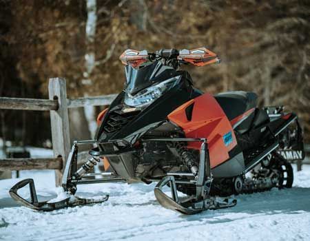 Black and orange snowmobile parked near a wooden fence in a snowy, wooded area.