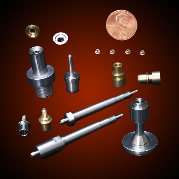 Assorted small metal machine parts, including tubes, pins, and a valve, next to a penny for scale.