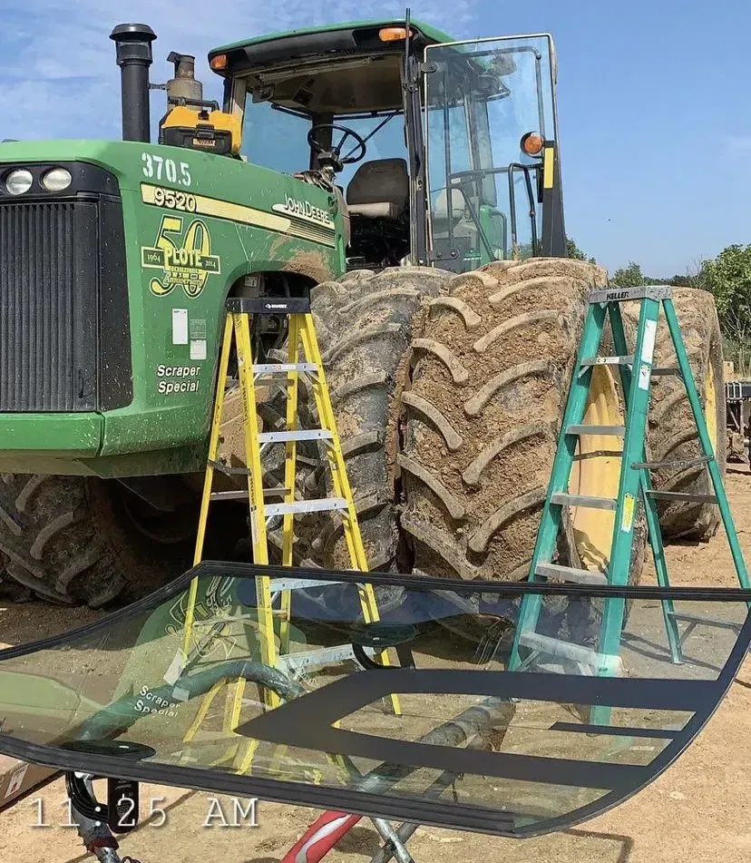 A John Deere tractor is parked outdoors near two ladders, with a large, rectangular glass window panel in the foreground.